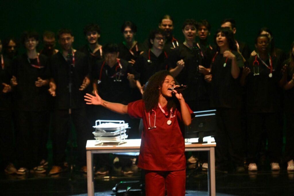 A performer in medical scrubs sings on stage during the opening of IncrEMentuM 2026, accompanied by a choir of clinicians under dramatic theatre lighting.