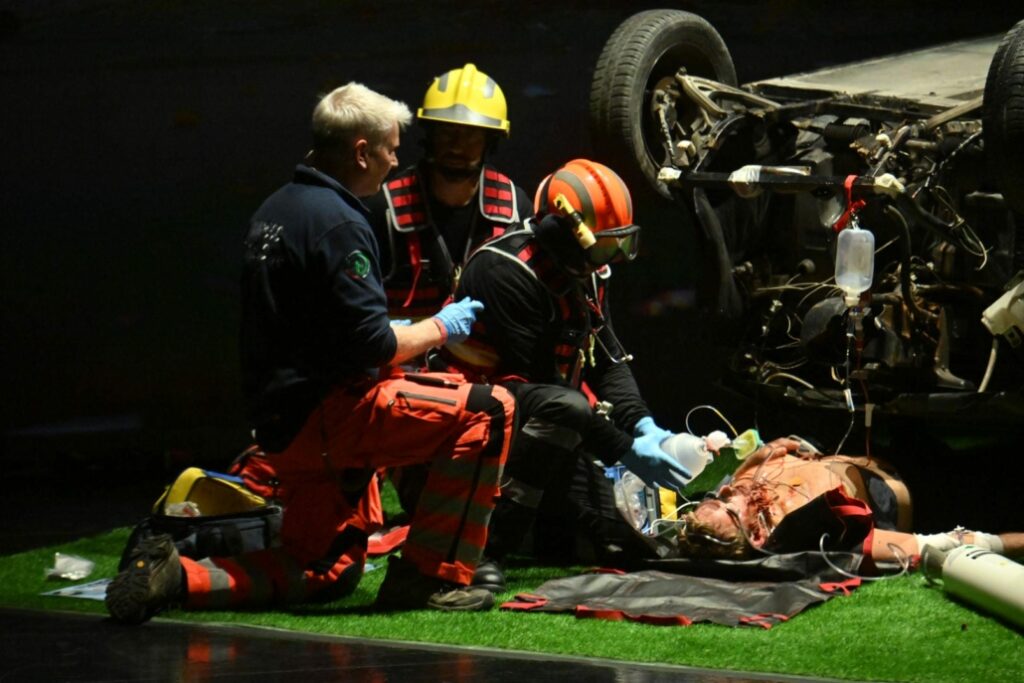 Emergency clinicians perform a live trauma simulation beside an overturned vehicle during IncrEMentuM 2026, demonstrating airway management and emergency care under stage lighting.