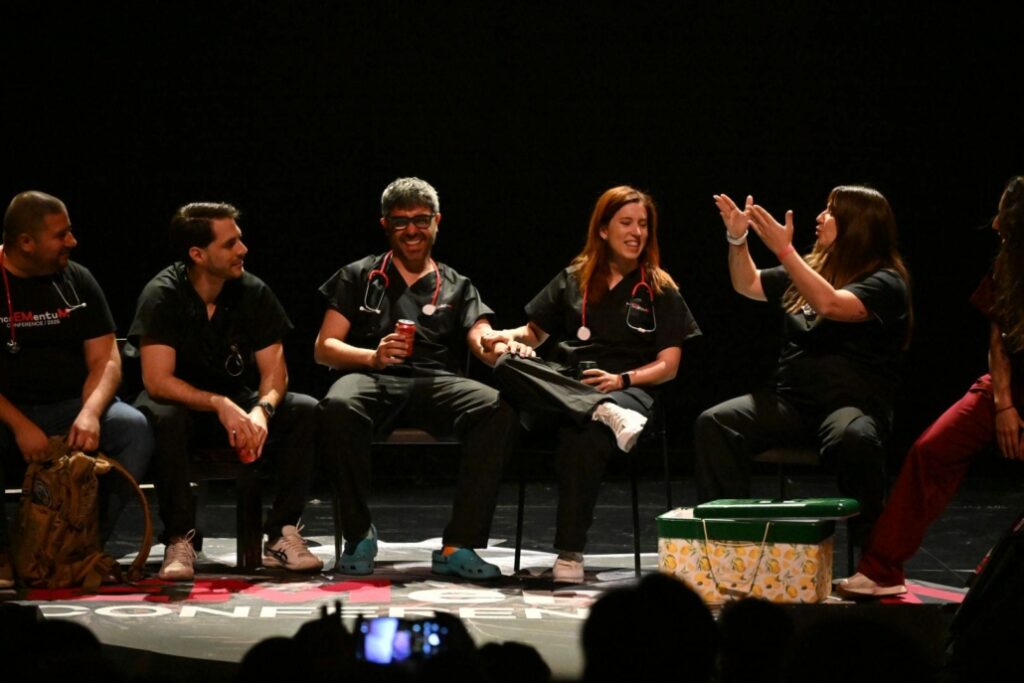 A group of clinicians sit on stage during a live simulation and discussion session at IncrEMentuM 2026, smiling and interacting under theatre lighting in front of a conference audience.