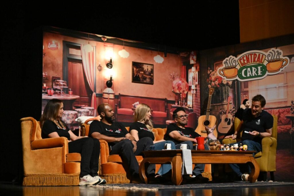 Speakers sit together on a Friends-inspired “Central Care” set during a hosted discussion at IncrEMentuM 2026, with sofas, coffee table props and informal conversation creating a relaxed conference atmosphere.