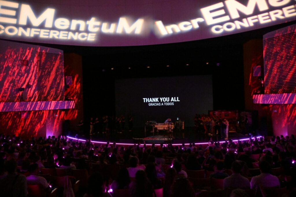 Audience members gather in a large auditorium during the closing moments of IncrEMentuM 2026 in Murcia, with illuminated conference branding surrounding the stage and a screen displaying “Thank You All / Gracias a Todos.”